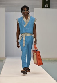 a man walking down a runway wearing a blue jumpsuit and orange bag